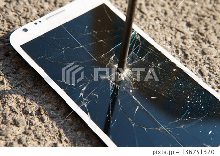 A white smartphone screen shattered by a metal spike on asphalt A white smartphone screen shattered by a metal spike on asphalt 136751320