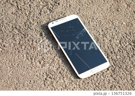 White smartphone with a severely cracked and shattered glass screen lying on rough outdoor asphalt 136751326