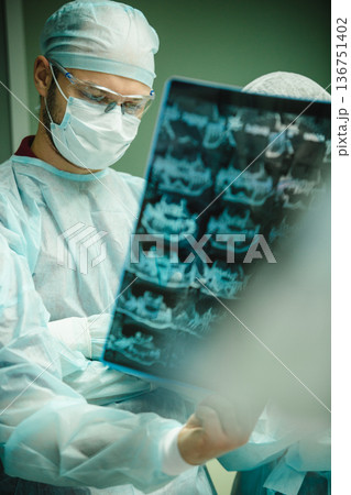 A focused male surgeon in sterile scrubs meticulously examines a dental X-ray A focused male surgeon in sterile scrubs meticulously examines a dental X-ray 136751402