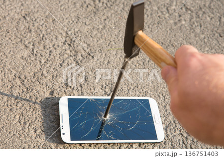 A hand drives a large metal nail through a smartphone screen with a hammer A hand drives a large metal nail through a smartphone screen with a hammer 136751403