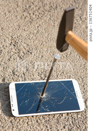 A hand drives a large metal nail through a smartphone screen with a hammer 136751404
