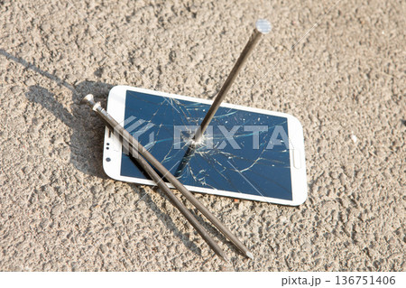 A white smartphone with a shattered screen pierced by a large metal nail on asphalt A white smartphone with a shattered screen pierced by a large metal nail on asphalt 136751406