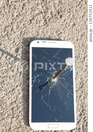 A white smartphone with a shattered screen pierced by a large metal nail on asphalt A white smartphone with a shattered screen pierced by a large metal nail on asphalt 136751411
