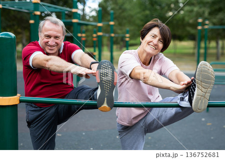 Mature couple doing leg stretching on crossbar together on sports ground of city park Mature couple doing leg stretching on crossbar together on sports ground of city park 136752681