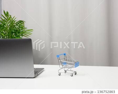 A metal toy shopping cart and an open gray laptop on a white table. 136752863