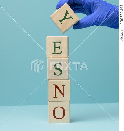 A hand in a blue medical glove places a wooden cube with the letter Y on a vertical stack of cubes, forming the word YES. 136752865