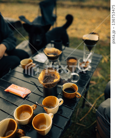 Friends enjoying morning pour-over coffee by campfire at rural campsite in mountains during sunrise 136753475