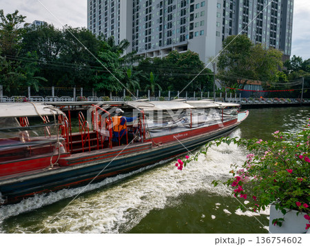 Passenger boat moving on canal water in city with green trees and flowers on bank, public water transportation system and commuting travel. 136754602