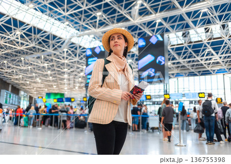 Bottom view of Caucasian young woman in straw hat smiling and holds passport. Defocused people in background. The concept of vacation and flight trip 136755398