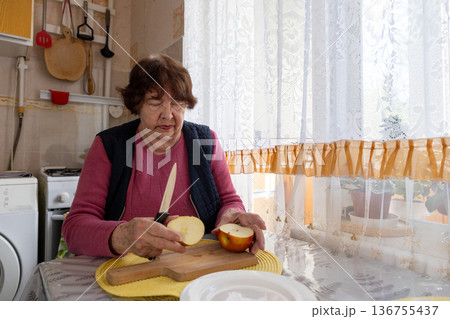 Elderly Caucasian woman cuts ripe apple on a kitchen table during daily meal preparation. She focuses on healthy eating and active aging. Concept of senior diet, prevention, and long life. Elderly Caucasian woman cuts ripe apple on a kitchen table during daily meal preparation. She focuses on healthy eating and active aging. Concept of senior diet, prevention, and long life. 136755437