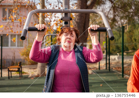 Elderly Caucasian woman trains on street gym machines during post surgery recovery. Sport helps restore mobility, health, and independence in later life. Elderly Caucasian woman trains on street gym machines during post surgery recovery. Sport helps restore mobility, health, and independence in later life. 136755446