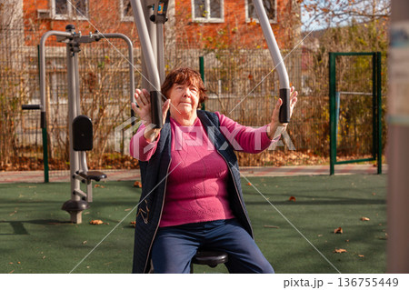 Elderly woman doing strength exercise on outdoor fitness equipment. Active lifestyle and senior wellness. Elderly woman doing strength exercise on outdoor fitness equipment. Active lifestyle and senior wellness. 136755449