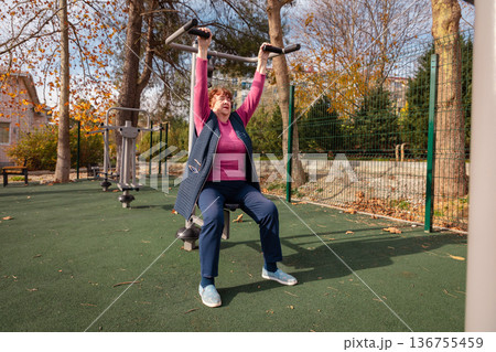 Older Caucasian woman using outdoor fitness machines in a green park. Physical activity, health, and long life in retirement. Older Caucasian woman using outdoor fitness machines in a green park. Physical activity, health, and long life in retirement. 136755459