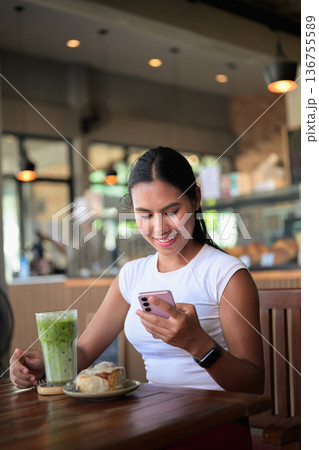 Smiling Woman Using Smartphone in Cafe with Green Iced Tea Matcha Latte Drink and Dessert 136755589