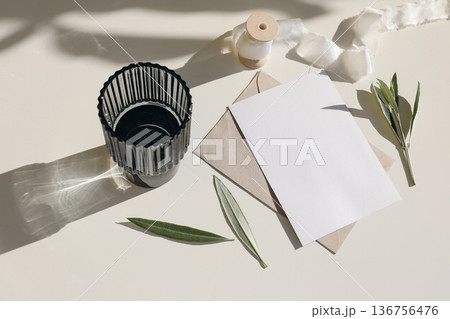 Minimalist stationery mockup. Blank greeting card, craft envelope. Olive leaves, ribbon spool. Glass of water on neutral background in sunlight, long shadows. Summer template in sunlight, copy space 136756476