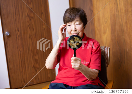 室内に座って、手鏡で顔を見ながらこめかみに手を当てるシニア女性 136757193
