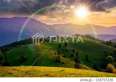 mountainous countryside scenery in springtime at sunset. dirt road and trees on the rolling hills in evening light. mountain ridge in the distance. alpine rural landscape. weather with rainbow 136757236