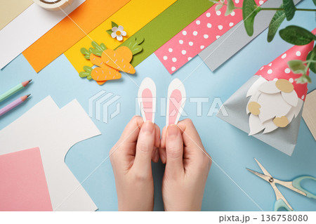 Hands hold paper rabbit ears. Colorful paper, scissors, tape on blue background. DIY handmade art project for kids. 136758080