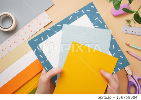 Hands holding colored paper for crafts. Tape, scissors, pencil on beige background. DIY handmade project for children. 136758084