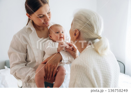 Standing, looking at each other. Mother, grandmother and little baby child on hands indoors 136758249