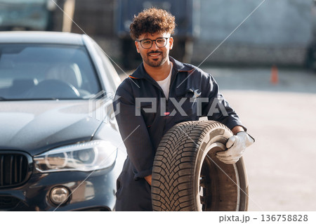 The tire in hands. Mechanic is with the car outdoors The tire in hands. Mechanic is with the car outdoors 136758828