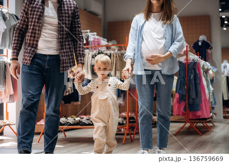 Walking and holding child by hands. Father, son and pregnant mother are in kid's clothing store Walking and holding child by hands. Father, son and pregnant mother are in kid's clothing store 136759669