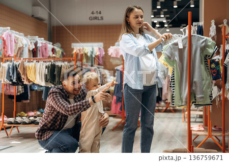 Showing what to buy. Father, son and pregnant mother are in kid's clothing store Showing what to buy. Father, son and pregnant mother are in kid's clothing store 136759671