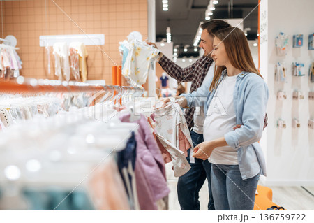 Clothes hanging in a row. Pregnant woman with man are in the kid's store 136759722