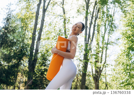 Orange colored yoga mat in hands. Beautiful fitness woman is outdoors, amazing nature Orange colored yoga mat in hands. Beautiful fitness woman is outdoors, amazing nature 136760338