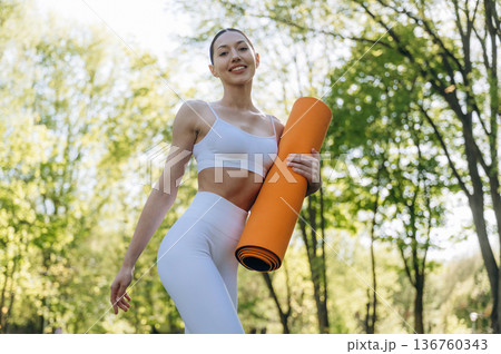 Orange colored yoga mat in hands. Beautiful fitness woman is outdoors, amazing nature 136760343
