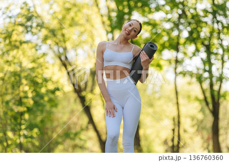 Front view. Standing with yoga mat. Beautiful fitness woman is outdoors, amazing nature 136760360