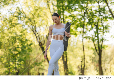 Front view. Standing with yoga mat. Beautiful fitness woman is outdoors, amazing nature Front view. Standing with yoga mat. Beautiful fitness woman is outdoors, amazing nature 136760362