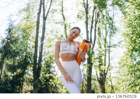 Orange colored yoga mat in hands. Beautiful fitness woman is outdoors, amazing nature Orange colored yoga mat in hands. Beautiful fitness woman is outdoors, amazing nature 136760529