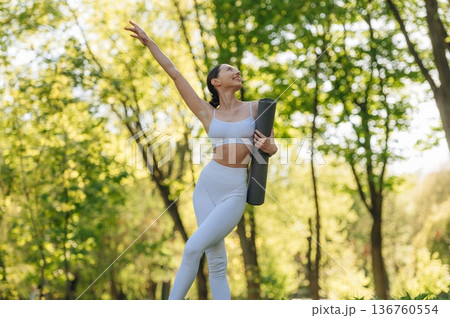 Front view. Standing with yoga mat. Beautiful fitness woman is outdoors, amazing nature Front view. Standing with yoga mat. Beautiful fitness woman is outdoors, amazing nature 136760554