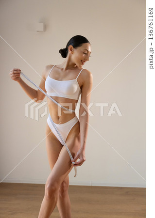 Hands are measuring the waist with length. Young woman in sportive clothes is against the wall Hands are measuring the waist with length. Young woman in sportive clothes is against the wall 136761169
