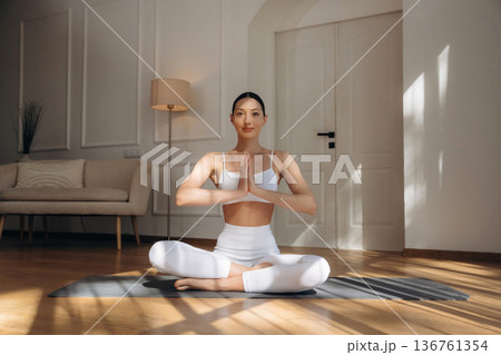 Making yoga gesture by hands, sitting in lotus pose. Young woman in fitness clothes is in the room Making yoga gesture by hands, sitting in lotus pose. Young woman in fitness clothes is in the room 136761354