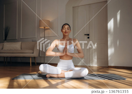 Making yoga gesture by hands, sitting in lotus pose. Young woman in fitness clothes is in the room Making yoga gesture by hands, sitting in lotus pose. Young woman in fitness clothes is in the room 136761355