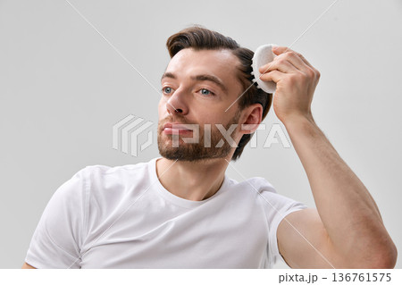 Adult short bearded man in white t shirt using silicone scalp brush on hair while looking forward, on light gray minimal studio background 136761575