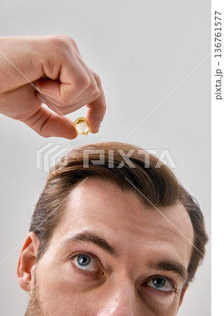 Close up of man applying liquid serum from capsule to hair while looking up on light gray background in minimal studio setting Close up of man applying liquid serum from capsule to hair while looking up on light gray background in minimal studio setting 136761577