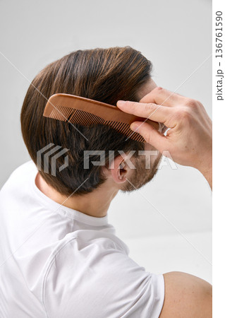 Elevated back angle shot of man in his thirties with short beard combing hair with wooden comb on light gray background 136761590