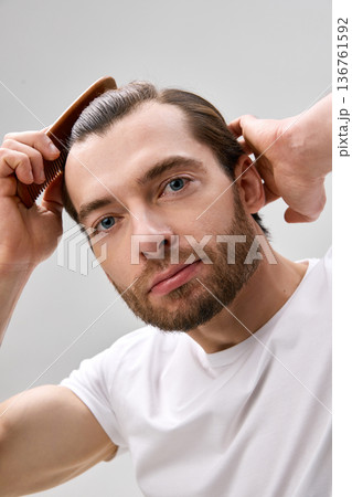 Close-up shot of man in his thirties with short beard combing hair with wooden comb on light gray background 136761592