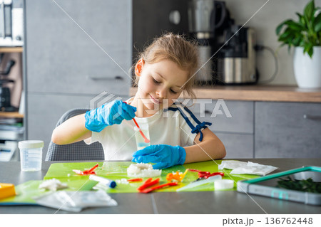 Young girl stirring chemical solution with measuring spoon in cup during crystal growing experiment at home. Chemistry practice and early STEM education through practical laboratory activity. 136762448