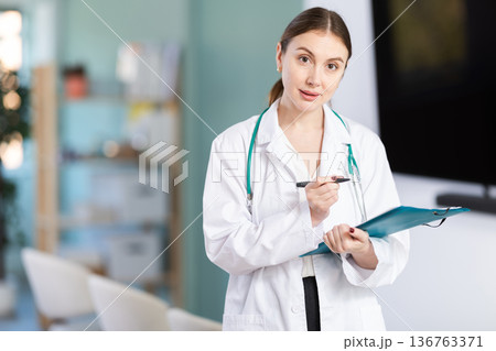 Portrait of young coach female doctor standing with folder with documents and materials in hands and talking in corporate seminar and inviting in auditorium 136763371