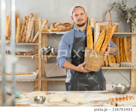 Male baker holds basket with finished products, shows many different baguettes 136763483