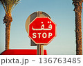 Red octagonal stop sign featuring Arabic and English text, positioned near palm trees under clear blue sky in outdoor environment 136763485