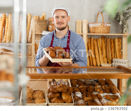 Man seller stands at bakery counter with basket filled with fresh croissants. 136763502