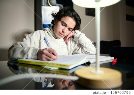 Teenage Boy Doing Homework At Home In The Evening Under A Desk Lamp Teenage Boy Doing Homework At Home In The Evening Under A Desk Lamp 136763799