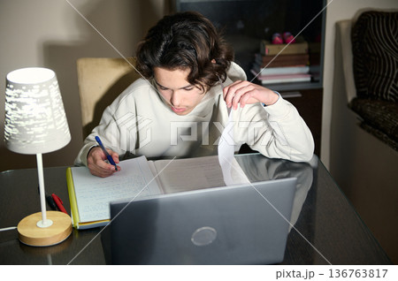 Teenage Boy Studying Homework At Home In The Evening With Laptop And Lamp Teenage Boy Studying Homework At Home In The Evening With Laptop And Lamp 136763817