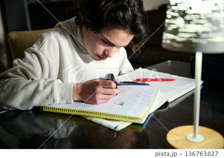 Teenage Boy Doing Homework At Night In Cozy Home Interior With Desk Lamp 136763827
