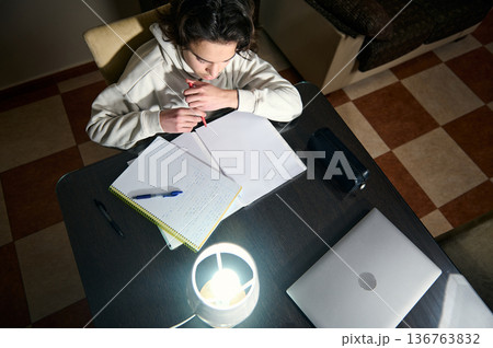 Teenage Boy Doing Homework At A Cozy Home Desk Under Warm Lamp Light In The Evening 136763832